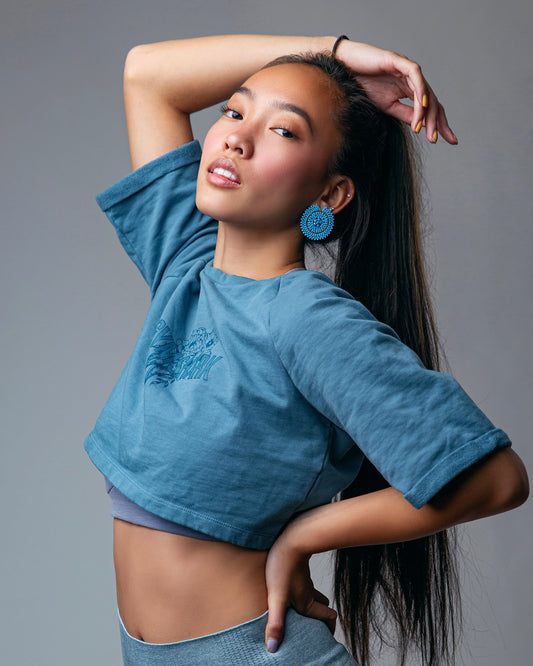 Woman wearing a blue stud earring, crop top and pants against a gray background
