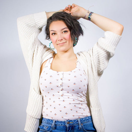 A person with short dark hair poses with both arms raised above their head, wearing Kawami turquoise hoop earrings, a white knit cardigan, a white floral-patterned tank top, and blue jeans, standing against a plain light-gray background