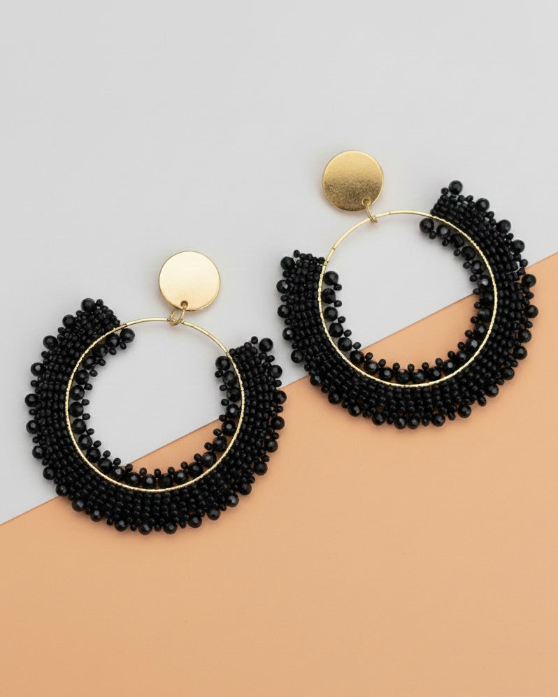 Black beaded hoop earrings with gold accents on a beige and gray background