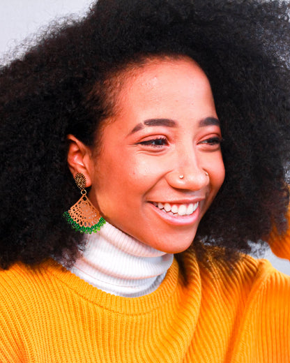 Woman with an afro wearing a bright orange sweater with green and gold Kawami earrings against a plain background