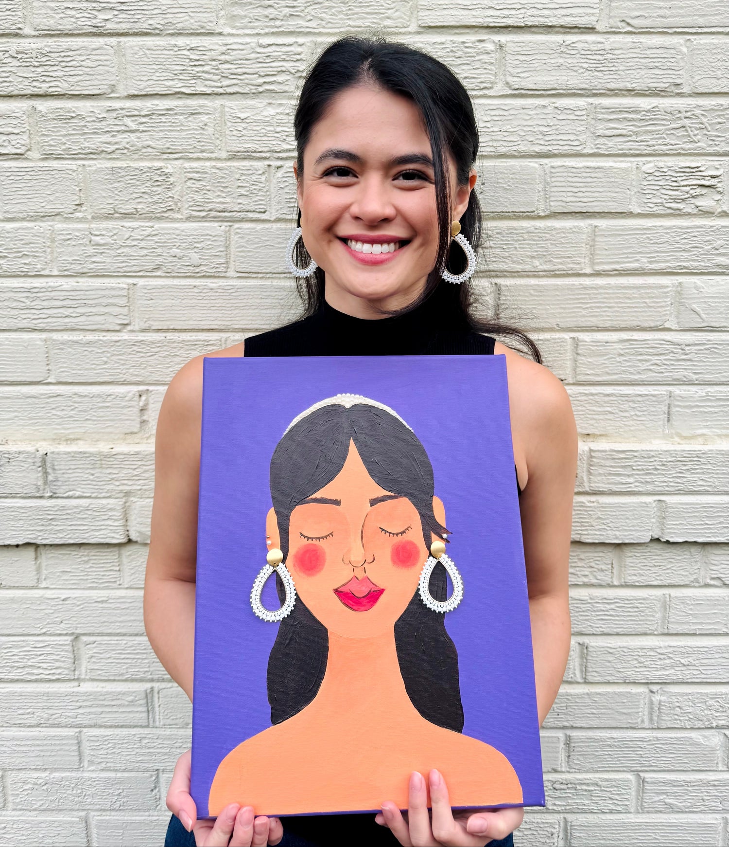 Woman holding a painting of a woman's face against a brick wall.
