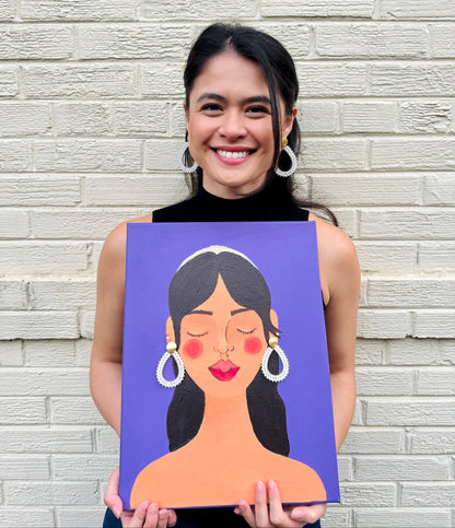 Woman holding a painting of a woman's face against a brick wall.