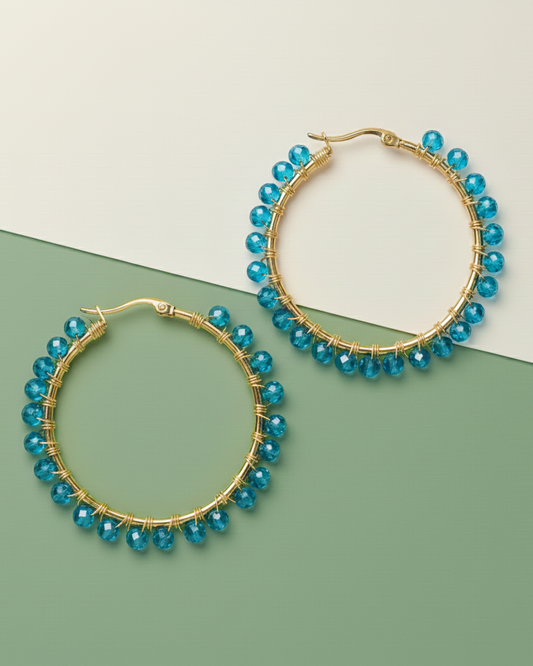 Gold hoop Kawami earrings with blue beads on a green and beige background