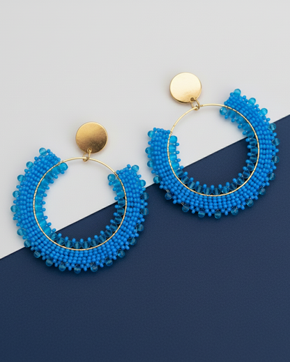 Blue beaded hoop Kawami earrings with gold accents on a two-tone blue background