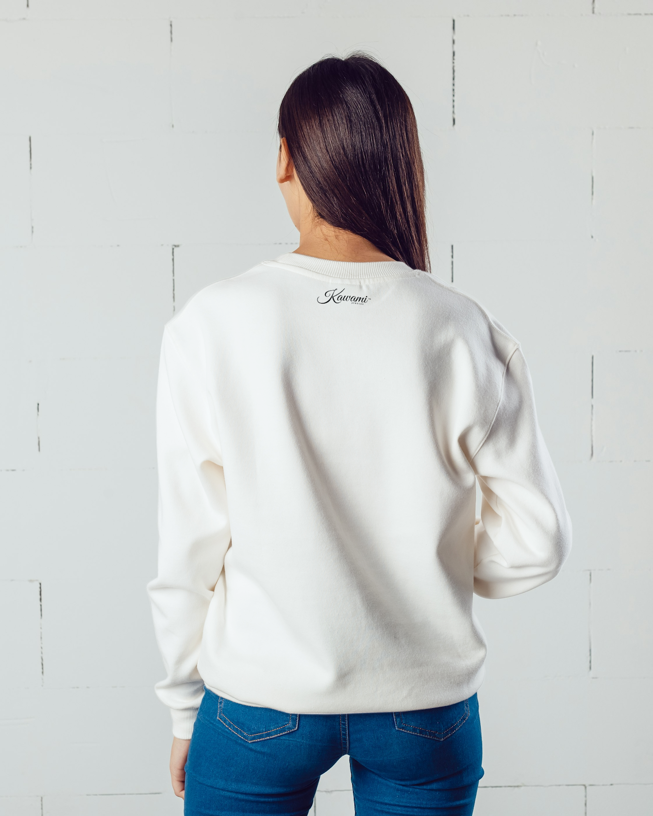 Person wearing a white sweatshirt with a brand logo on the back, standing against a light gray brick wall.