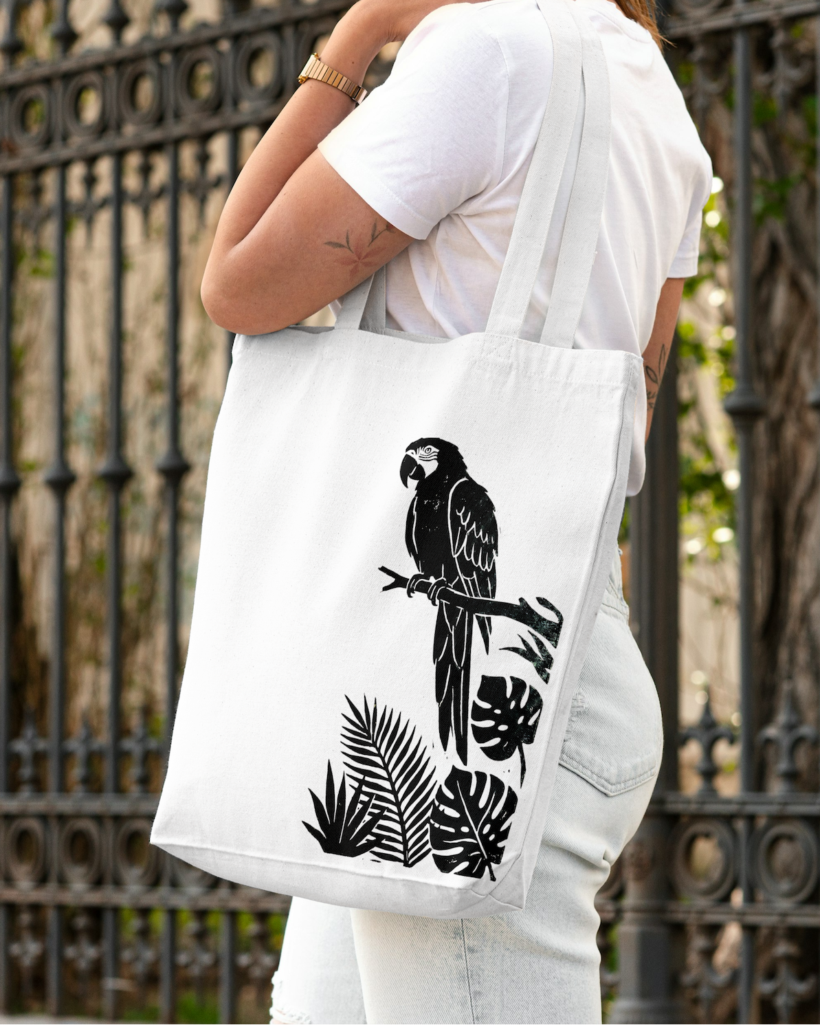 Person holding a white tote bag with black parrot design against an iron fence background