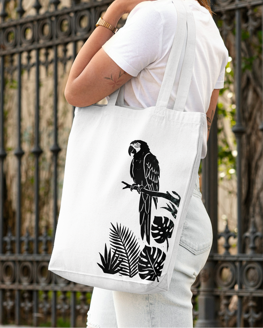 Person holding a white tote bag with black parrot design against an iron fence background