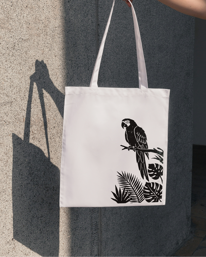 White tote bag with black parrot design held by a hand against a gray wall.