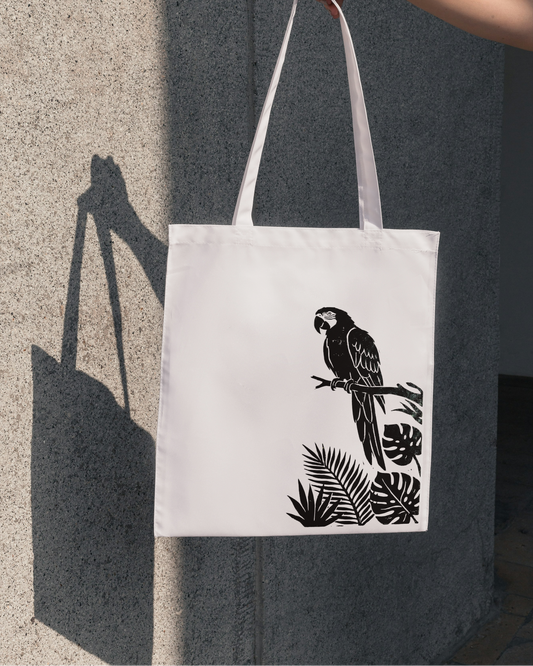 White tote bag with black parrot design held by a hand against a gray wall.