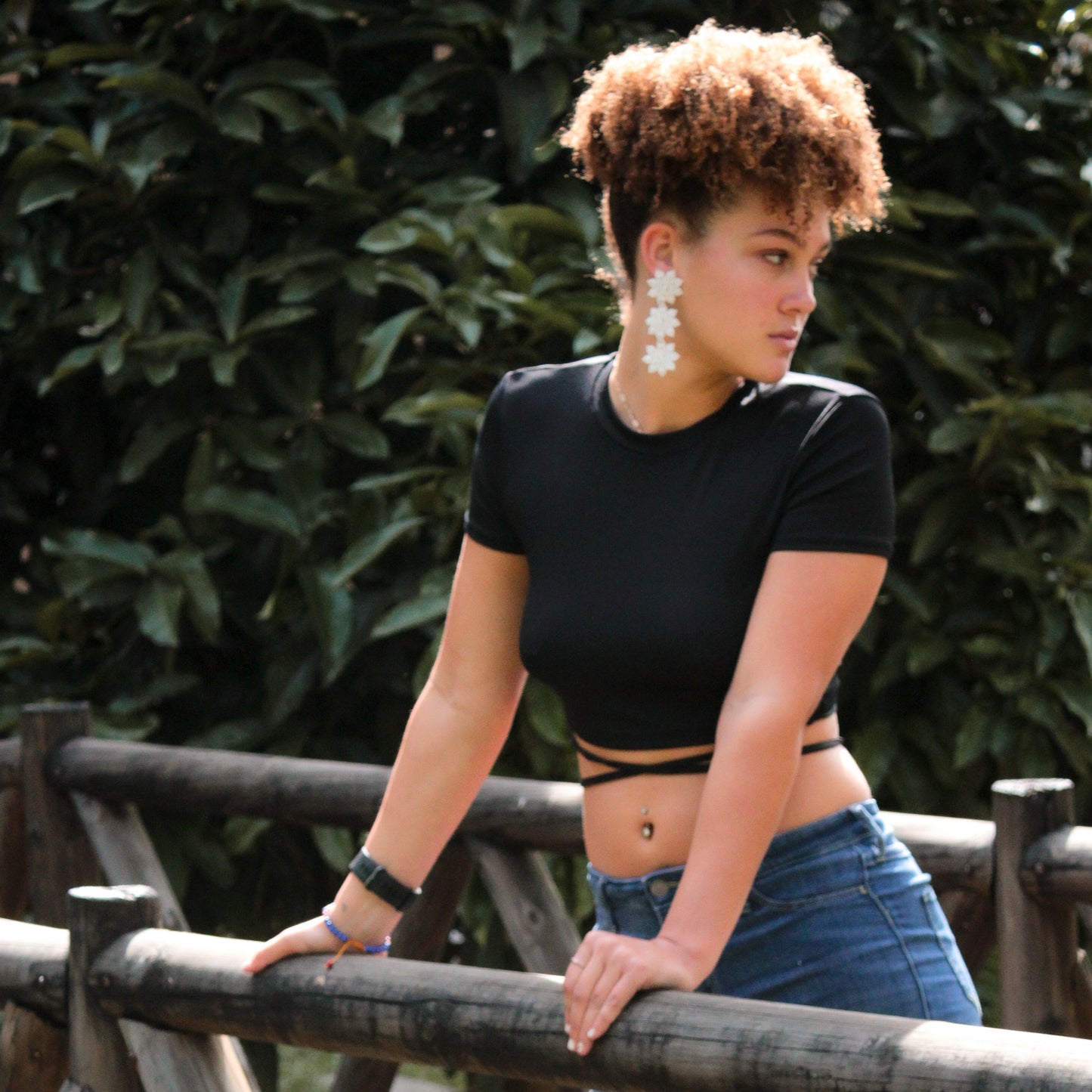 A woman with curly hair pulled up into a puff leans against a wooden fence outdoors, wearing a black crop top with crisscross straps, blue jeans, and large white floral-shaped earrings. She looks to the side with a neutral expression, with greenery in the background