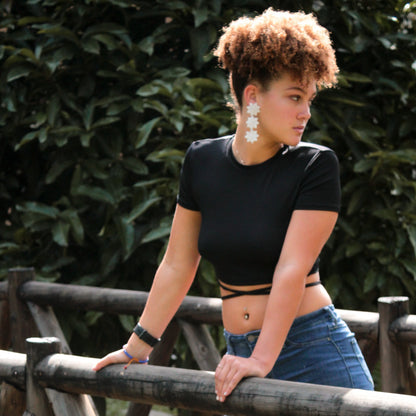 A woman with curly hair pulled up into a puff leans against a wooden fence outdoors, wearing a black crop top with crisscross straps, blue jeans, and large white floral-shaped earrings. She looks to the side with a neutral expression, with greenery in the background