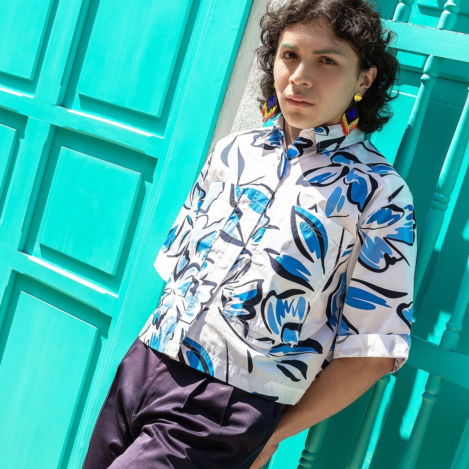 Person wearing a blue-and-white patterned shirt and colorful beaded earrings, standing in sunlight against a bright turquoise door and wall